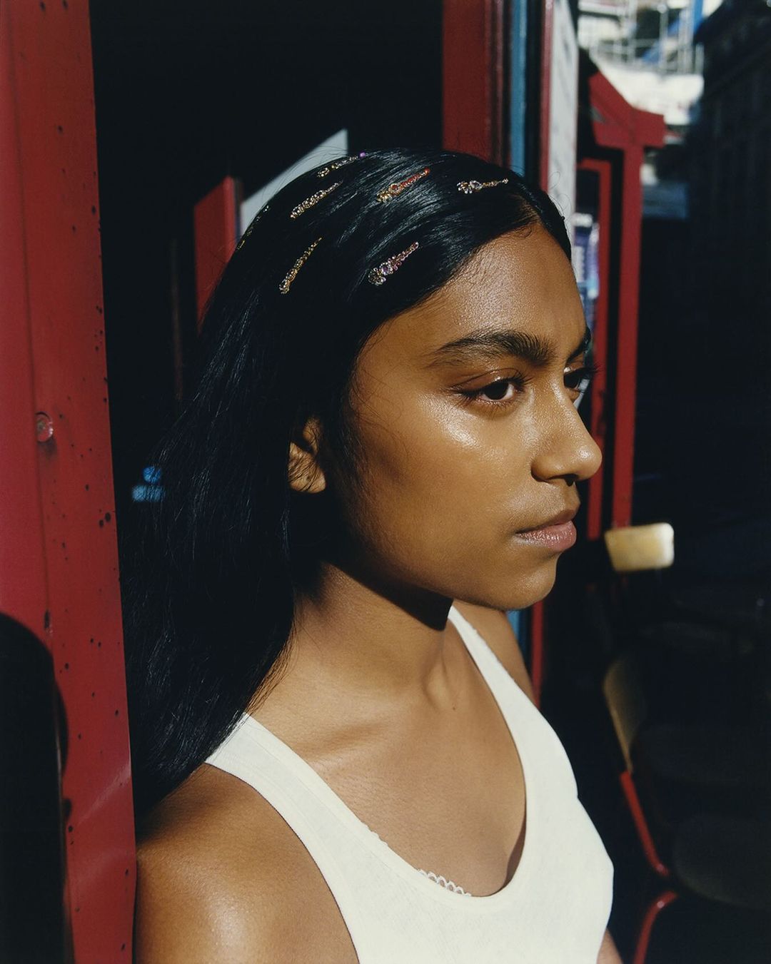 Portrait of a young woman with dark hair wearing a white top and hair clips, standing in front of a red and blue structure.
