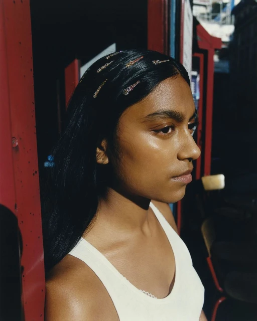 Portrait of a young woman with dark hair wearing a white top and hair clips, standing in front of a red and blue structure.