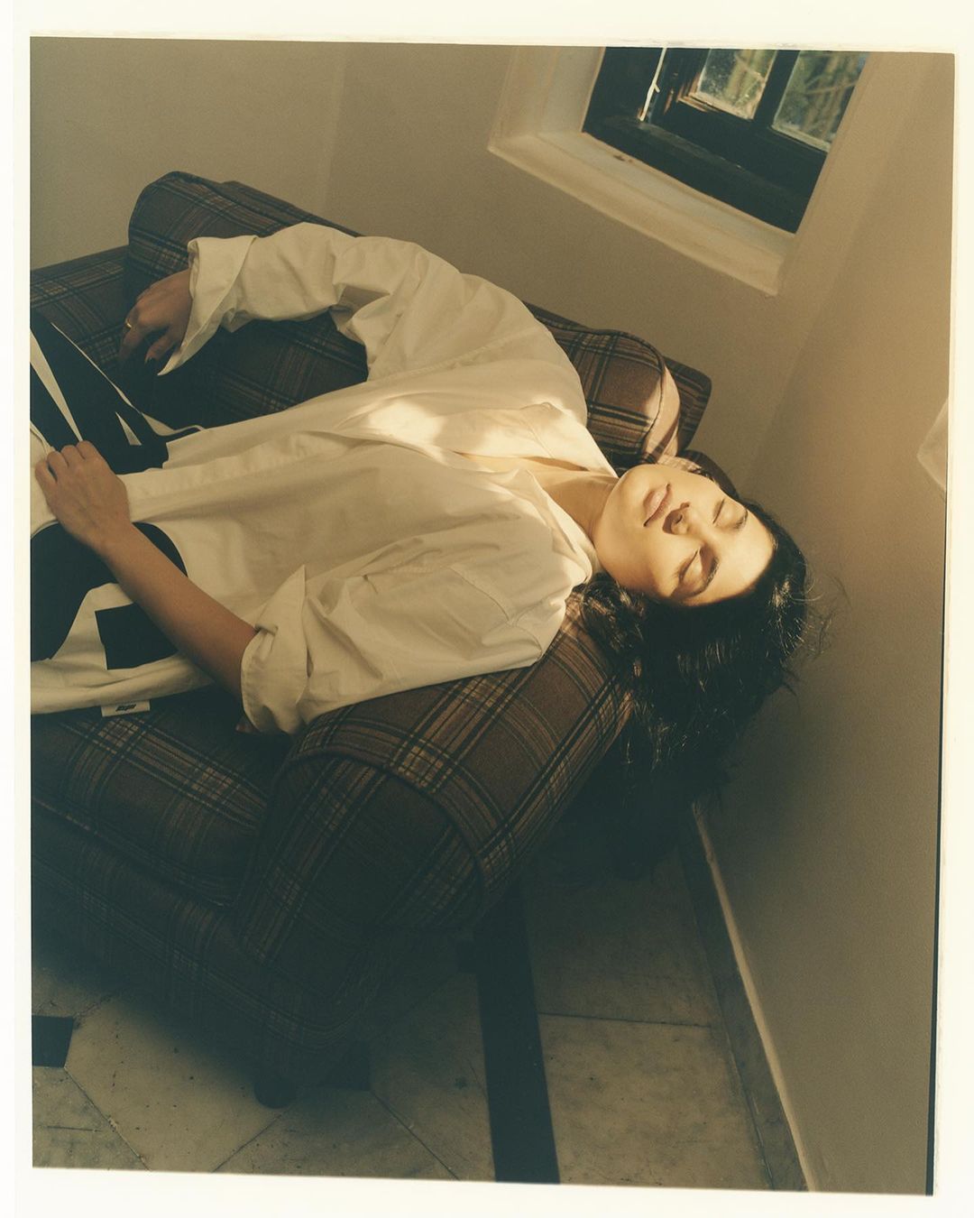 Woman in white shirt lying on plaid chair in interior. Dark curly hair, relaxed expression, natural light, soft shadows.