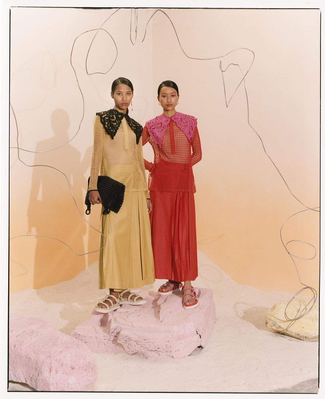 Two Asian fashion models wearing matching outfits with oversized collars and platform sandals stand on a pink rock in a studio setting.