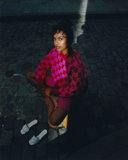 Woman sits cross-legged on cobblestones in checkerboard sweater and slides, highlighted by sunlight, creating a fashionable outdoor portrait.
