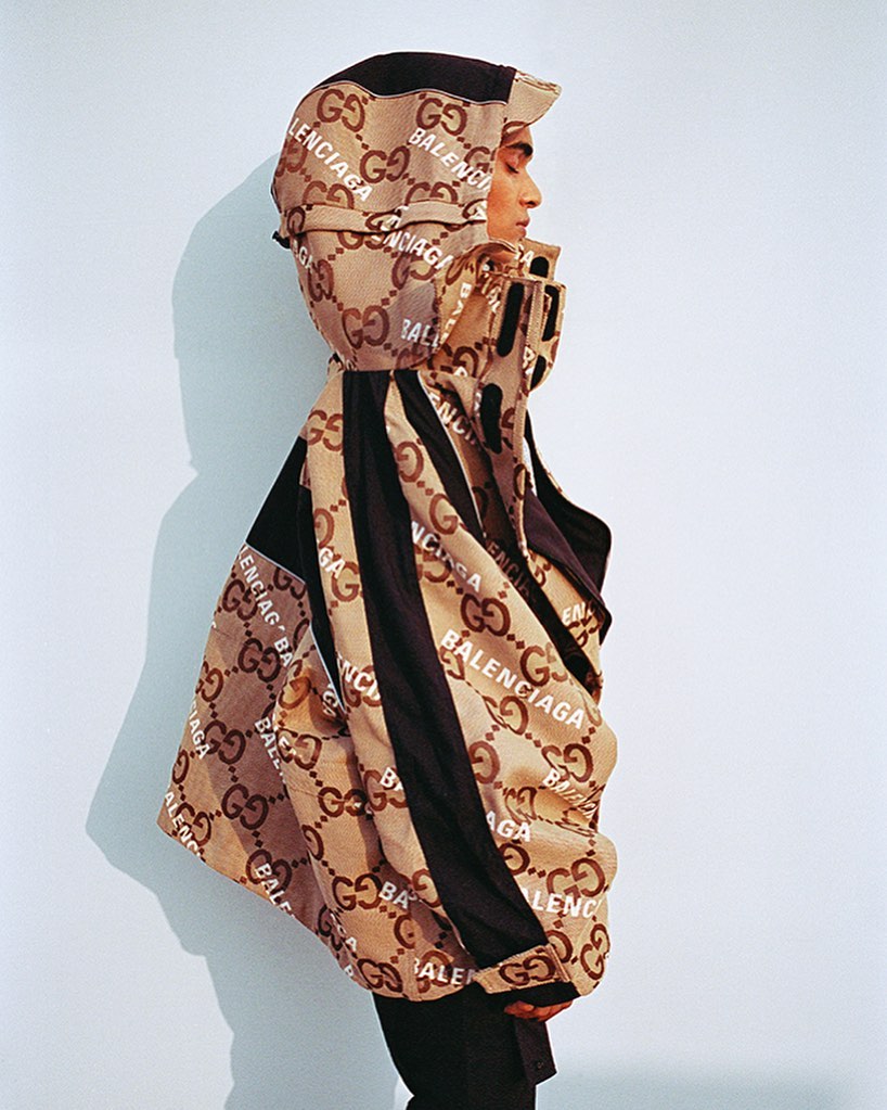 Side profile studio shot of a young adult wearing a beige and brown jacket with the logos of Gucci and Balenciaga all over it.
