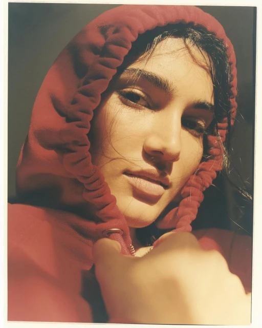 Close-up portrait of a woman with fair skin, wearing a red hooded sweatshirt, partially obscuring her face with the hood in soft lighting.