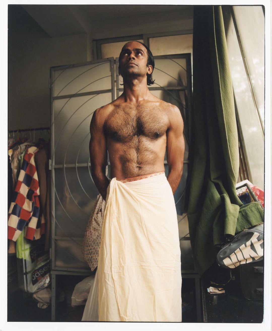 Man with dark hair, bare chest, and a white wrap stands in front of a metal cabinet looking up, lit by window light.