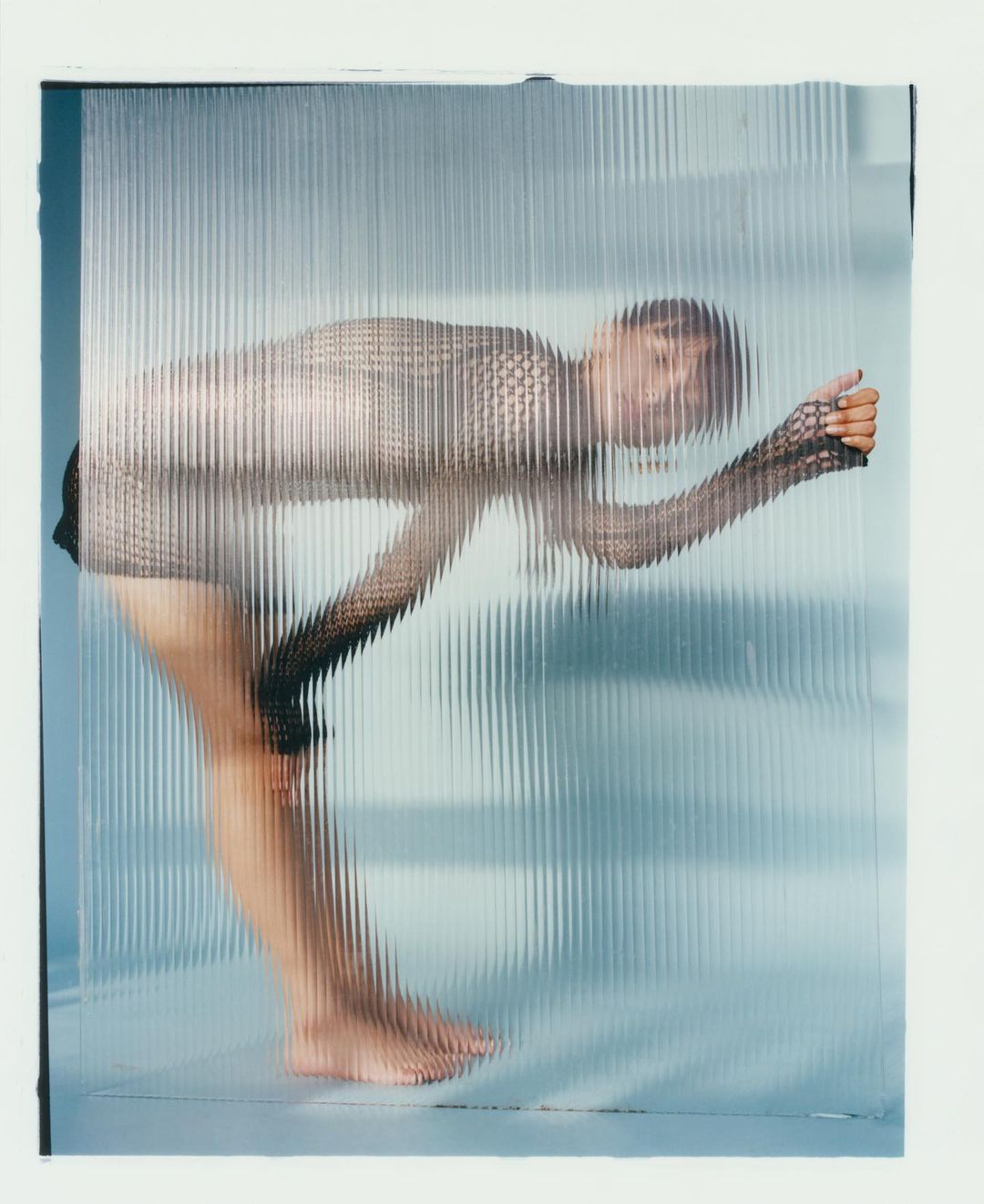 Fashionable woman in a mesh top stands behind a textured glass panel in a brightly lit studio setting, creating a distorted effect.