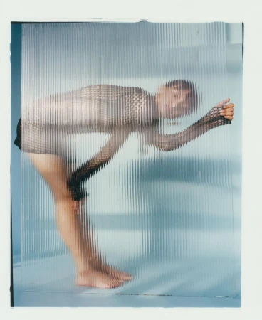 Fashionable woman in a mesh top stands behind a textured glass panel in a brightly lit studio setting, creating a distorted effect.