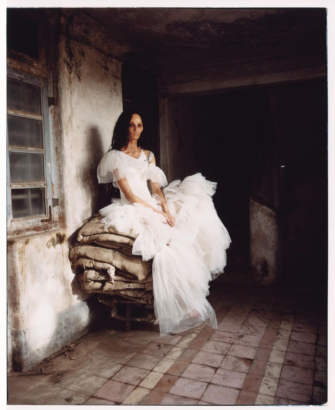 Elegant woman in a white dress sits on stacked burlap sacks inside a weathered building with tile floors and dim light, creating a moody scene.
