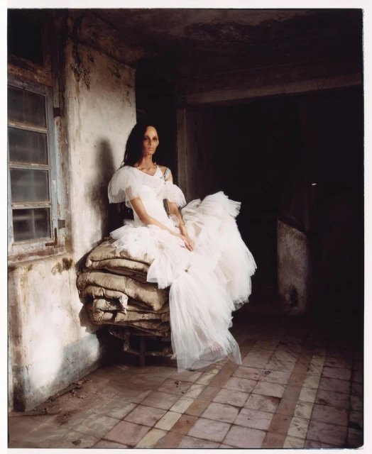Elegant woman in a white dress sits on stacked burlap sacks inside a weathered building with tile floors and dim light, creating a moody scene.
