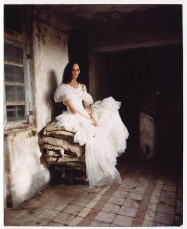 Elegant woman in a white dress sits on stacked burlap sacks inside a weathered building with tile floors and dim light, creating a moody scene.