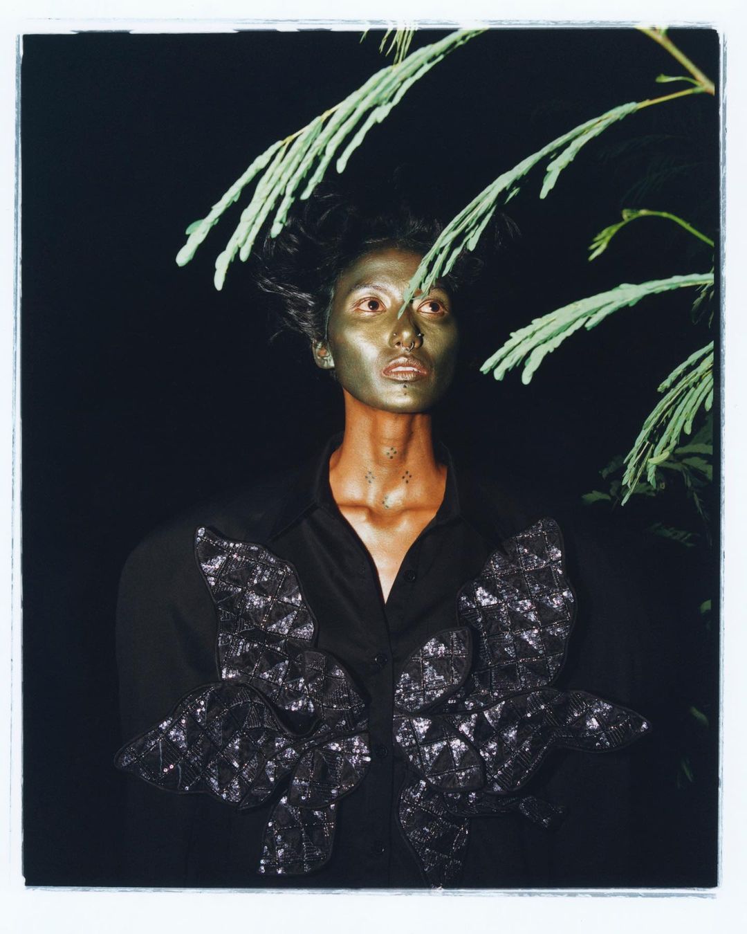 Eye-level shot of a person with metallic makeup, wearing a black shirt with decorative butterfly accents, positioned against a dark background.