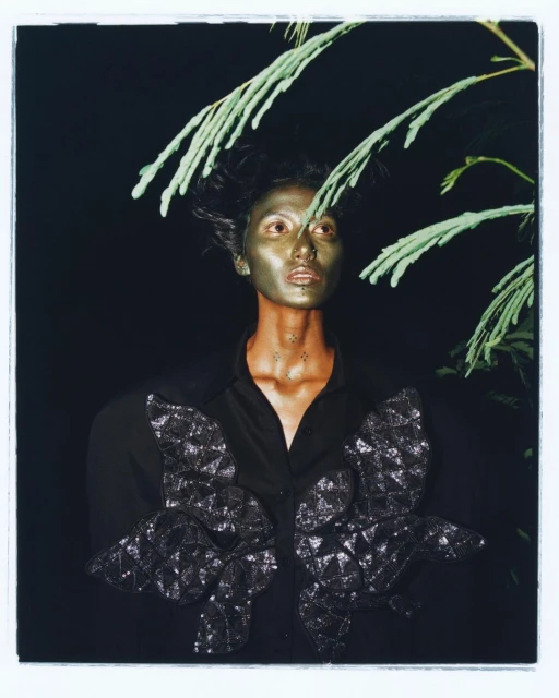 Eye-level shot of a person with metallic makeup, wearing a black shirt with decorative butterfly accents, positioned against a dark background.
