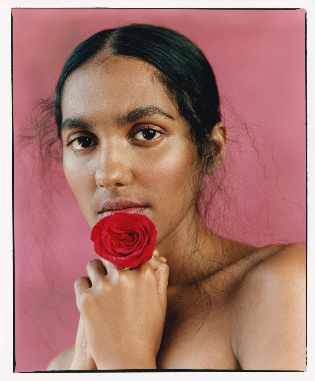 Close-up of a model with dark hair holding a red rose to her mouth against a pink background in a beauty or fashion photograph.