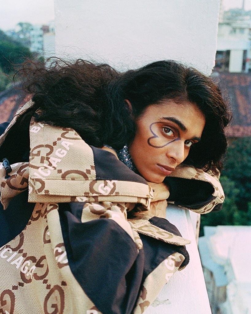 Portrait of a person with dark hair and a heart drawn around their eye wearing a beige and brown patterned jacket with an outdoor background.