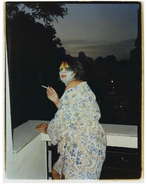 Woman with make-up and patterned robe poses on balcony, smoking a cigarette at dusk with dark cityscape in the background.