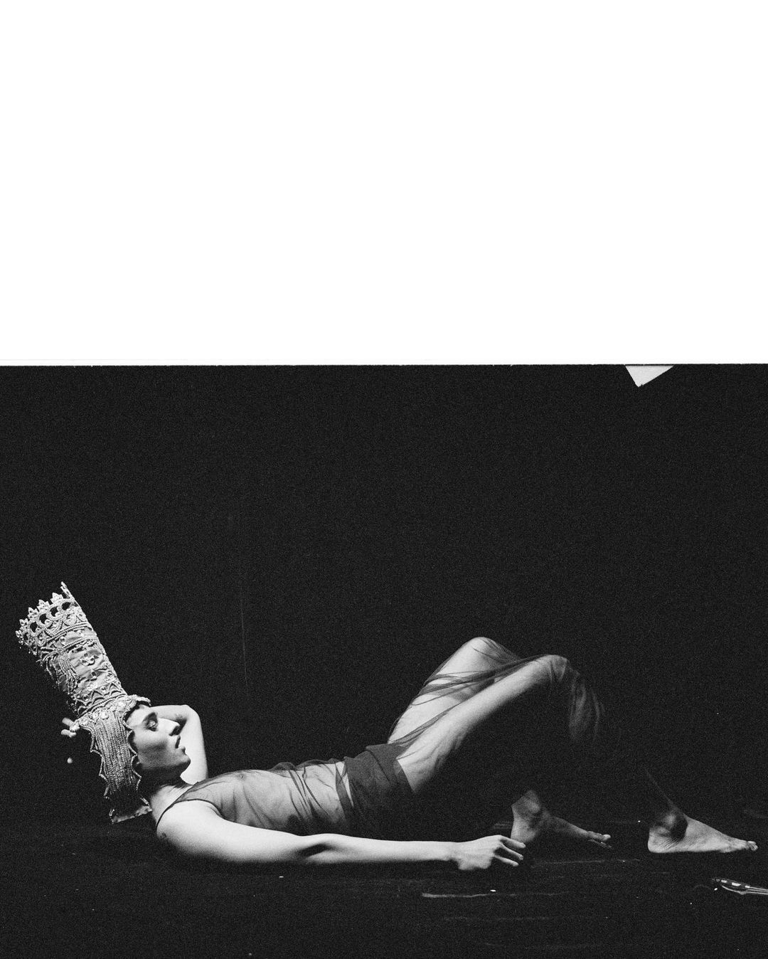 Woman with elaborate headpiece reclines on floor in a black-and-white studio shot, creating a striking visual with minimalist composition.
