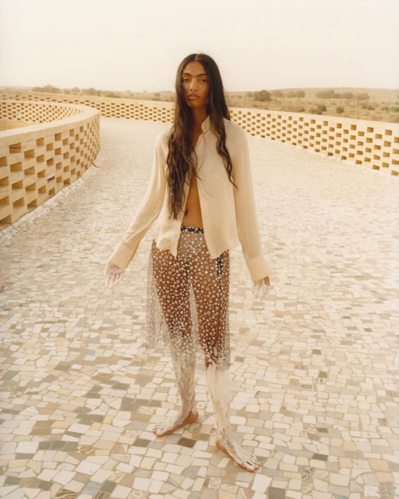 Fashion shot of a young adult woman with long hair, wearing an unbuttoned shirt and transparent skirt, posing on a tiled walkway.