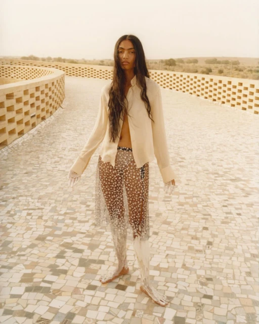 Fashion shot of a young adult woman with long hair, wearing an unbuttoned shirt and transparent skirt, posing on a tiled walkway.