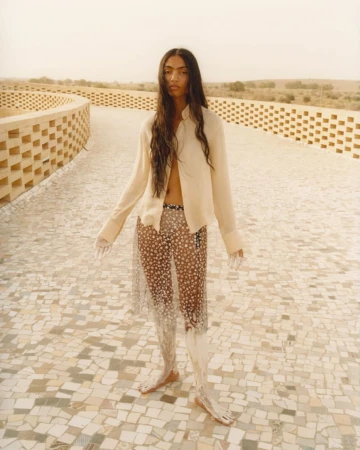 Fashion shot of a young adult woman with long hair, wearing an unbuttoned shirt and transparent skirt, posing on a tiled walkway.