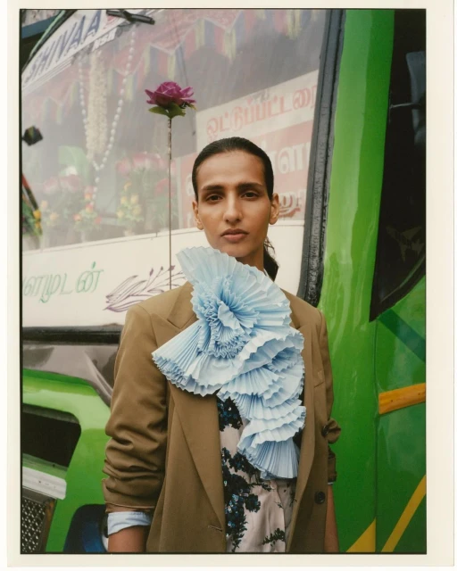 Portrait of a person wearing a tan blazer and a large light blue frill with a green bus and lettering in the background.