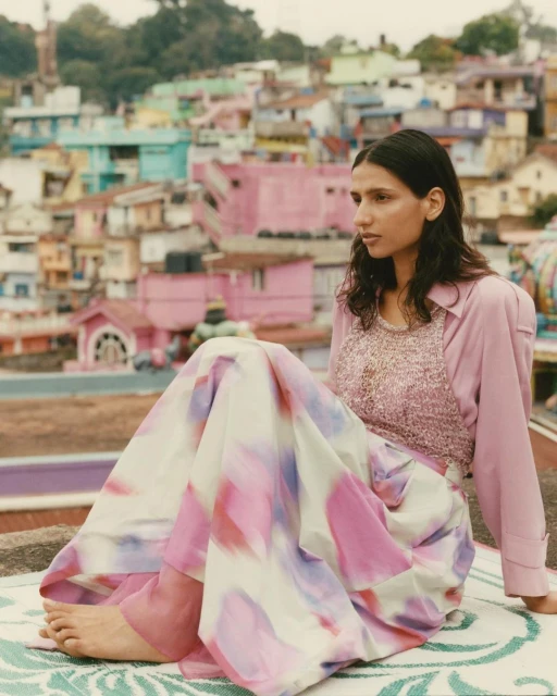 Adult female model wearing pink, white, and purple dress sitting outdoors on a patterned rug in front of an urban village scene.
