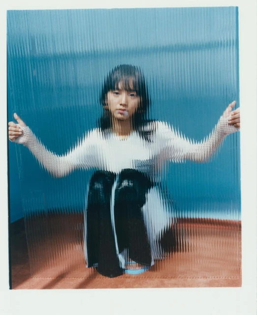 Young woman with dark hair poses behind textured glass in a medium shot, wearing white t-shirt and dark pants, sitting on a reddish surface.