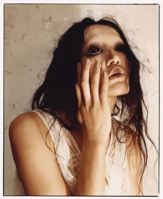 Headshot of a woman with dark hair and dramatic makeup in a white dress, with her hand near her face against a textured wall.