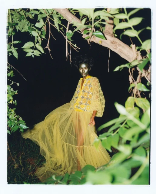 A woman with gold body paint wears a yellow dress surrounded by green foliage in a dark, artistic outdoor fashion portrait.
