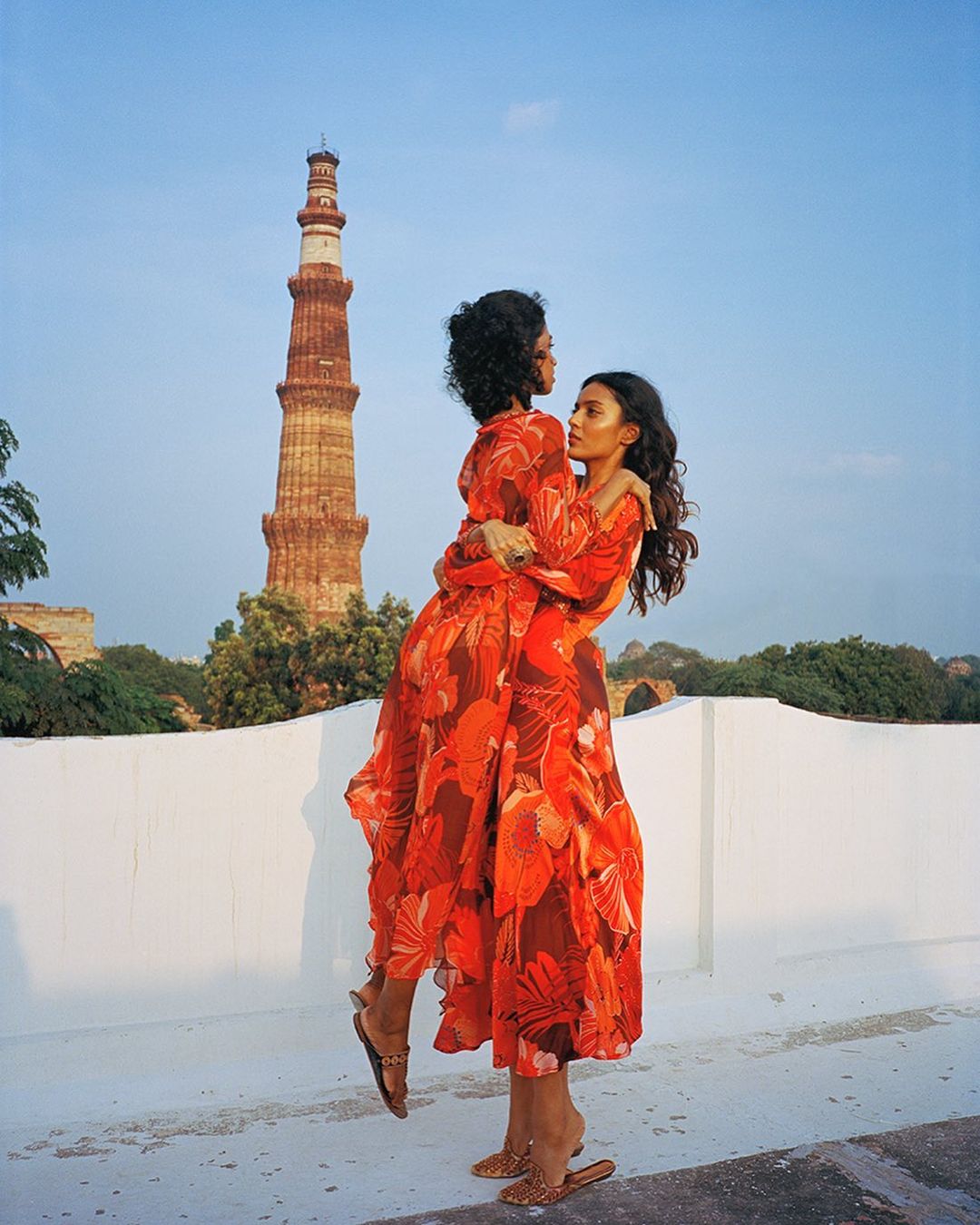 Two adult females embrace in orange floral dresses with the Qutub Minar visible in the background on a rooftop on a sunny day.