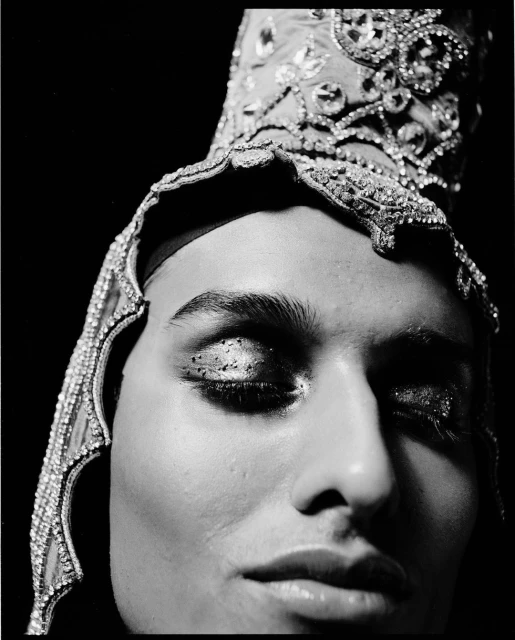 Striking black and white close-up of a person with glitter eyeshadow and an elaborate, jeweled headpiece against a dark backdrop.