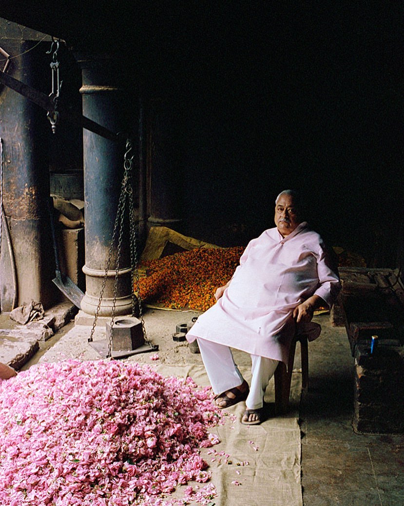 An elderly man sits on a stool amid pink and orange flowers in a dimly lit room with iron columns and overhead chains.