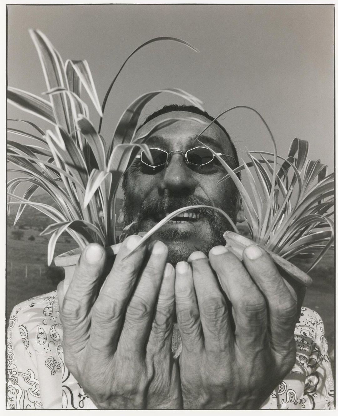 Monochrome portrait of a bearded man with glasses holding two potted spider plants, with a field visible in the soft-focus background.