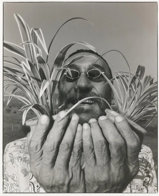Monochrome portrait of a bearded man with glasses holding two potted spider plants, with a field visible in the soft-focus background.