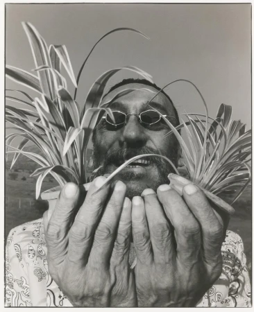 Monochrome portrait of a bearded man with glasses holding two potted spider plants, with a field visible in the soft-focus background.