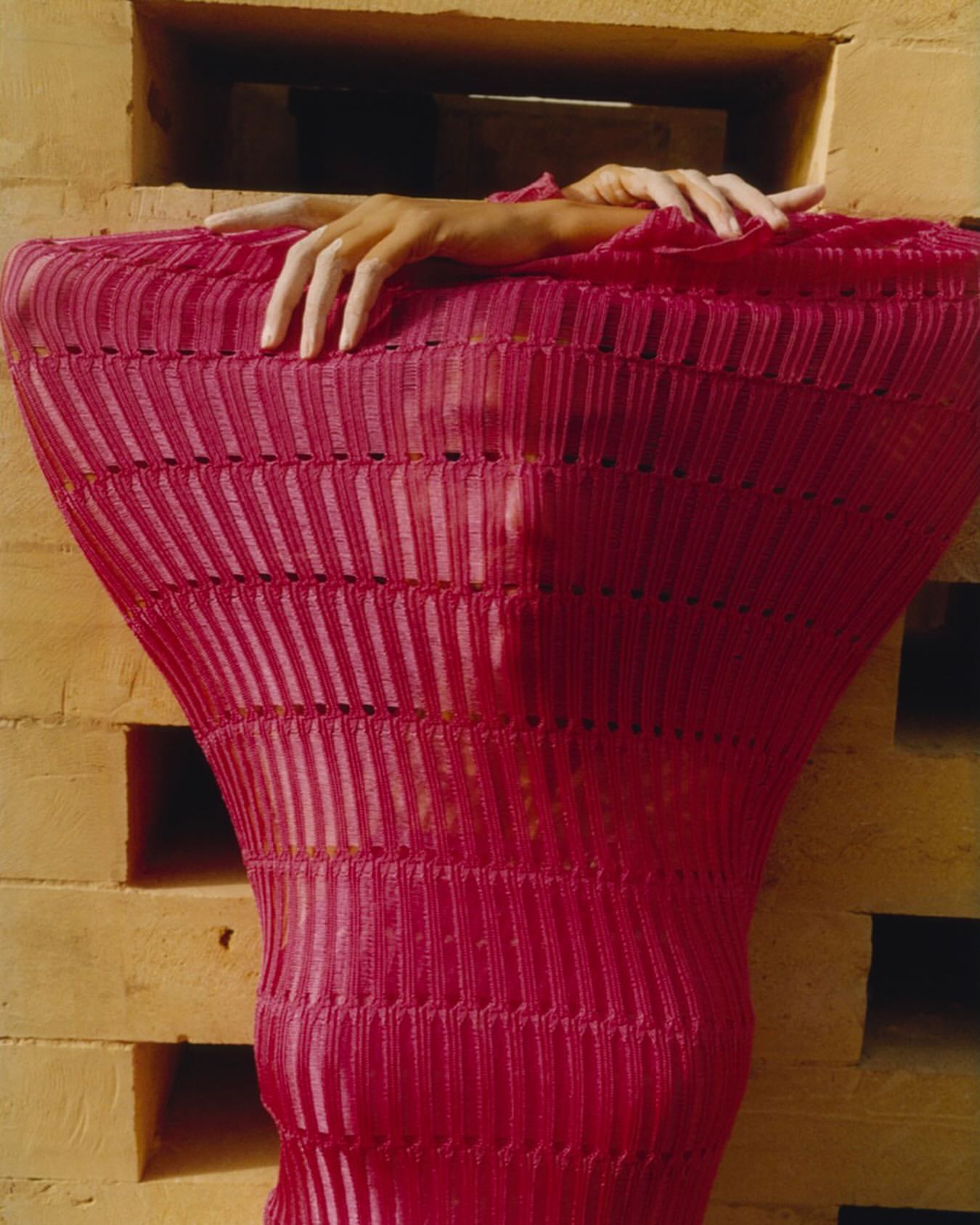 A person in a pink textured garment stands framed by tan brick blocks, creating an artistic fashion portrait with unique color and texture.