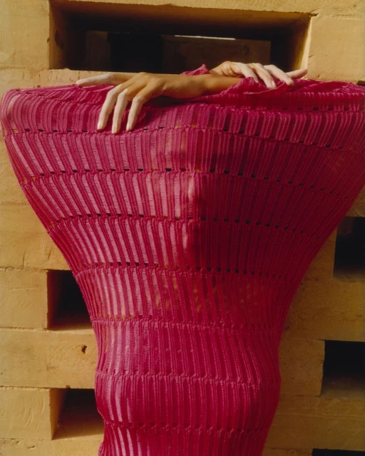 A person in a pink textured garment stands framed by tan brick blocks, creating an artistic fashion portrait with unique color and texture.