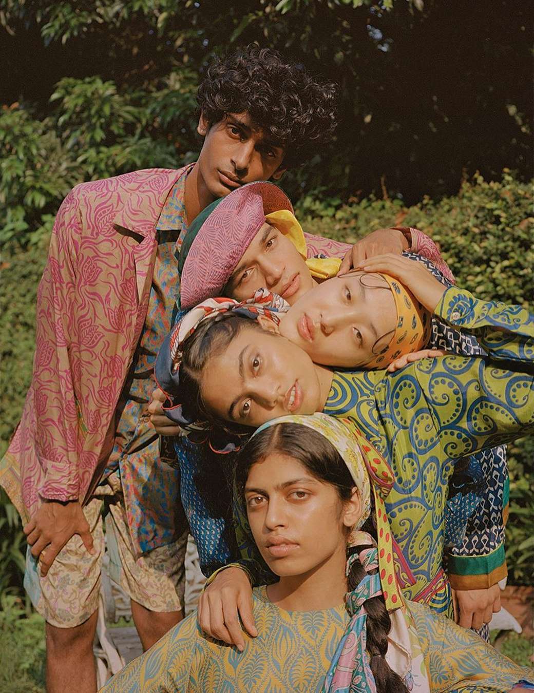 Group of four people in patterned clothing posing outdoors in front of green foliage. The image has a vintage, editorial feel.