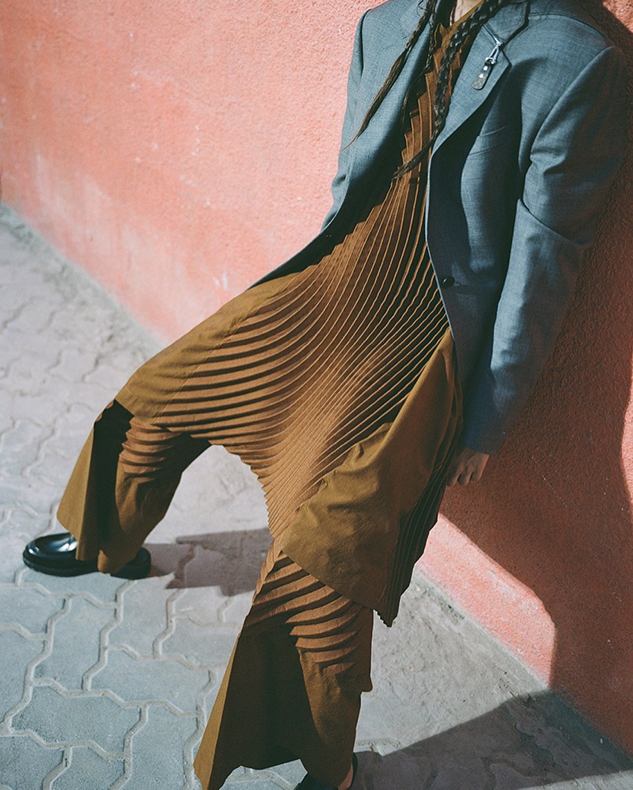 Person wearing pleated brown jumpsuit with a grey blazer standing against a peach-colored wall on a patterned grey stone surface.