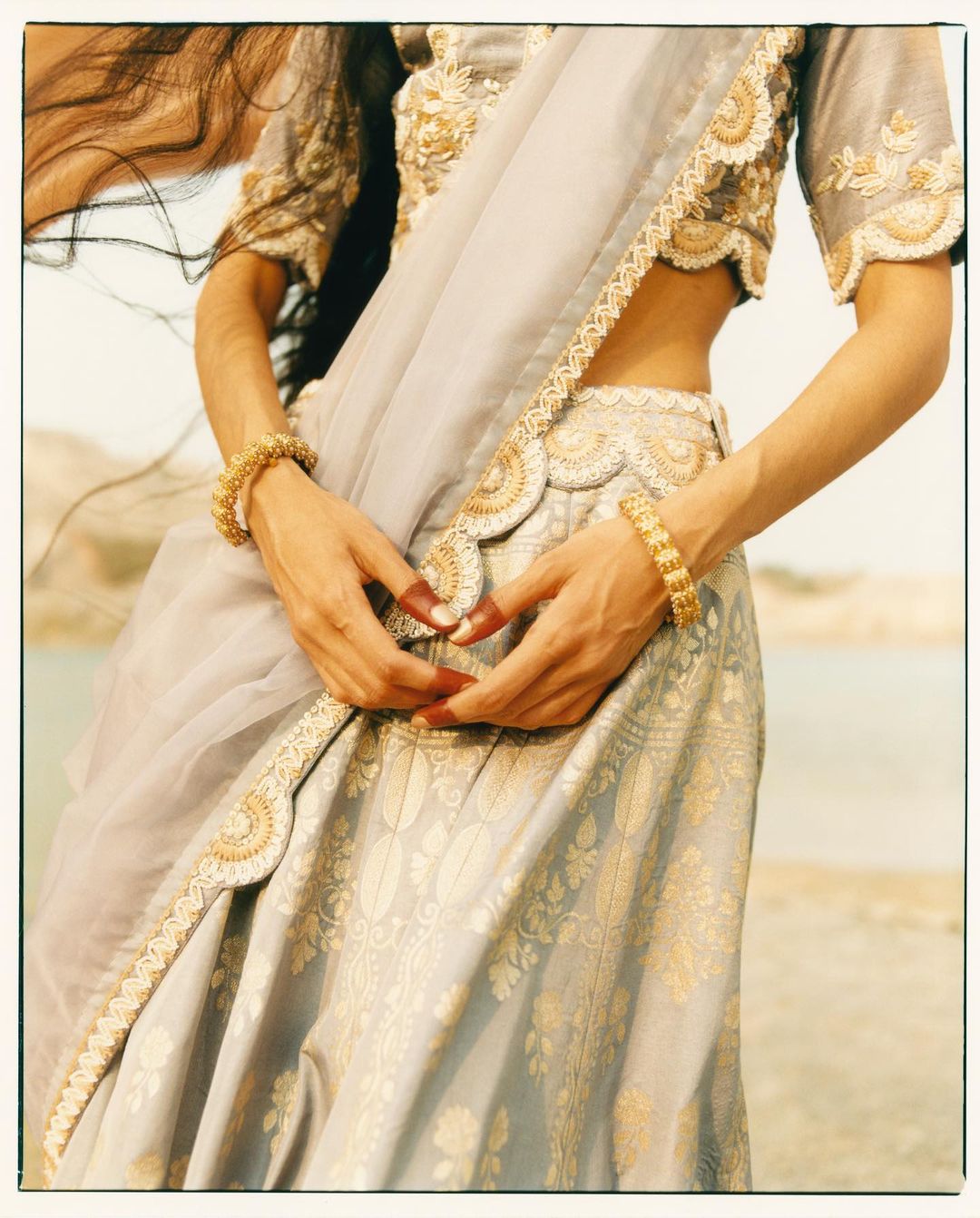 Model in traditional Indian dress featuring a saree and jewellery poses outside near the water with neutral-toned sand and sky.