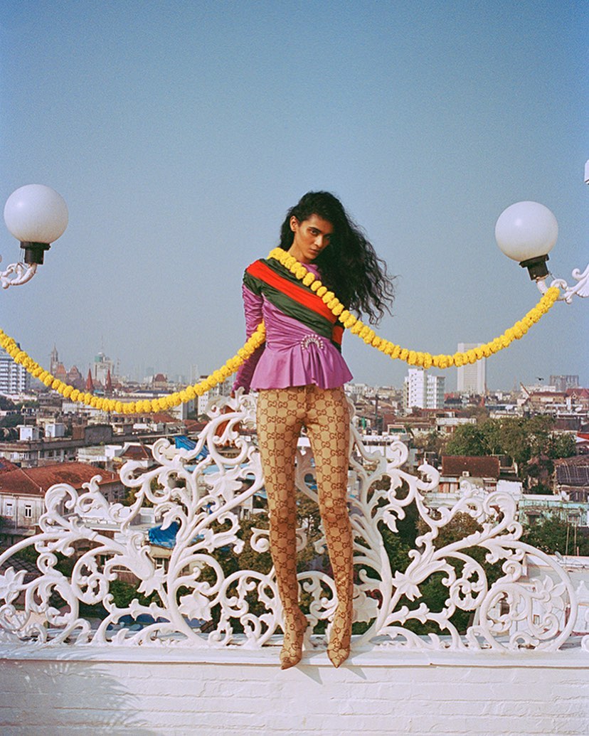 Fashion shot of a slender woman wearing a lavender top and monogram tights, standing on a rooftop with an urban skyline behind her.