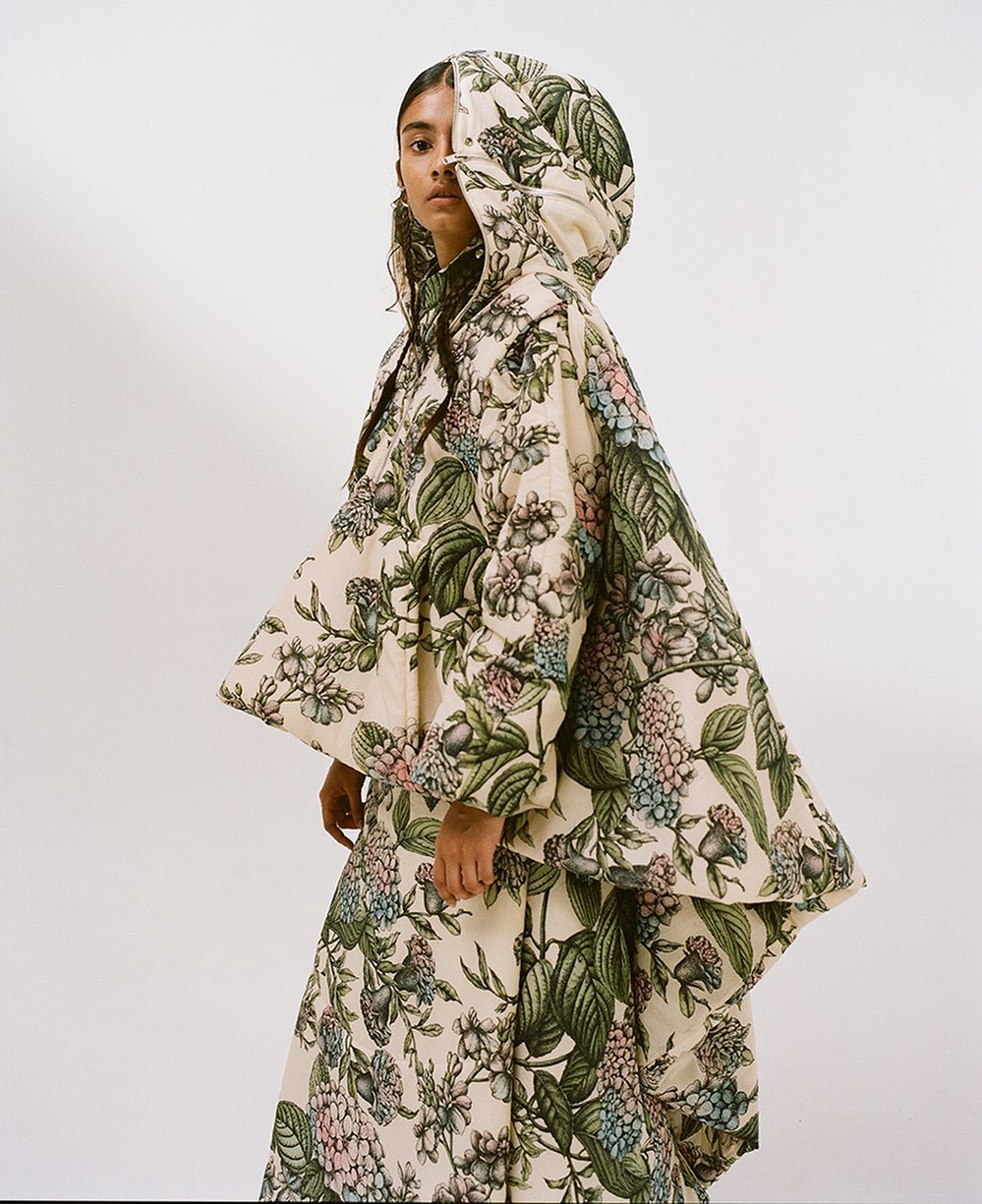 A woman in a hooded poncho and matching skirt with a floral pattern poses against a white background in a fashion editorial shot.