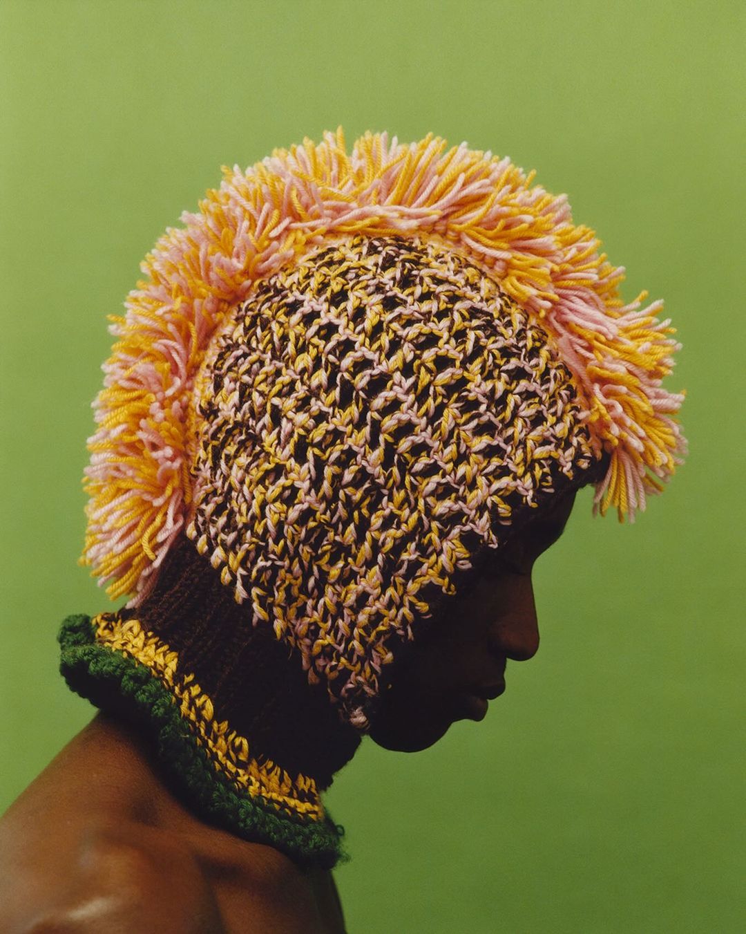 Person in profile wearing a knitted mohawk hat with yellow and pink faux fur accents against a green background in a studio setting.