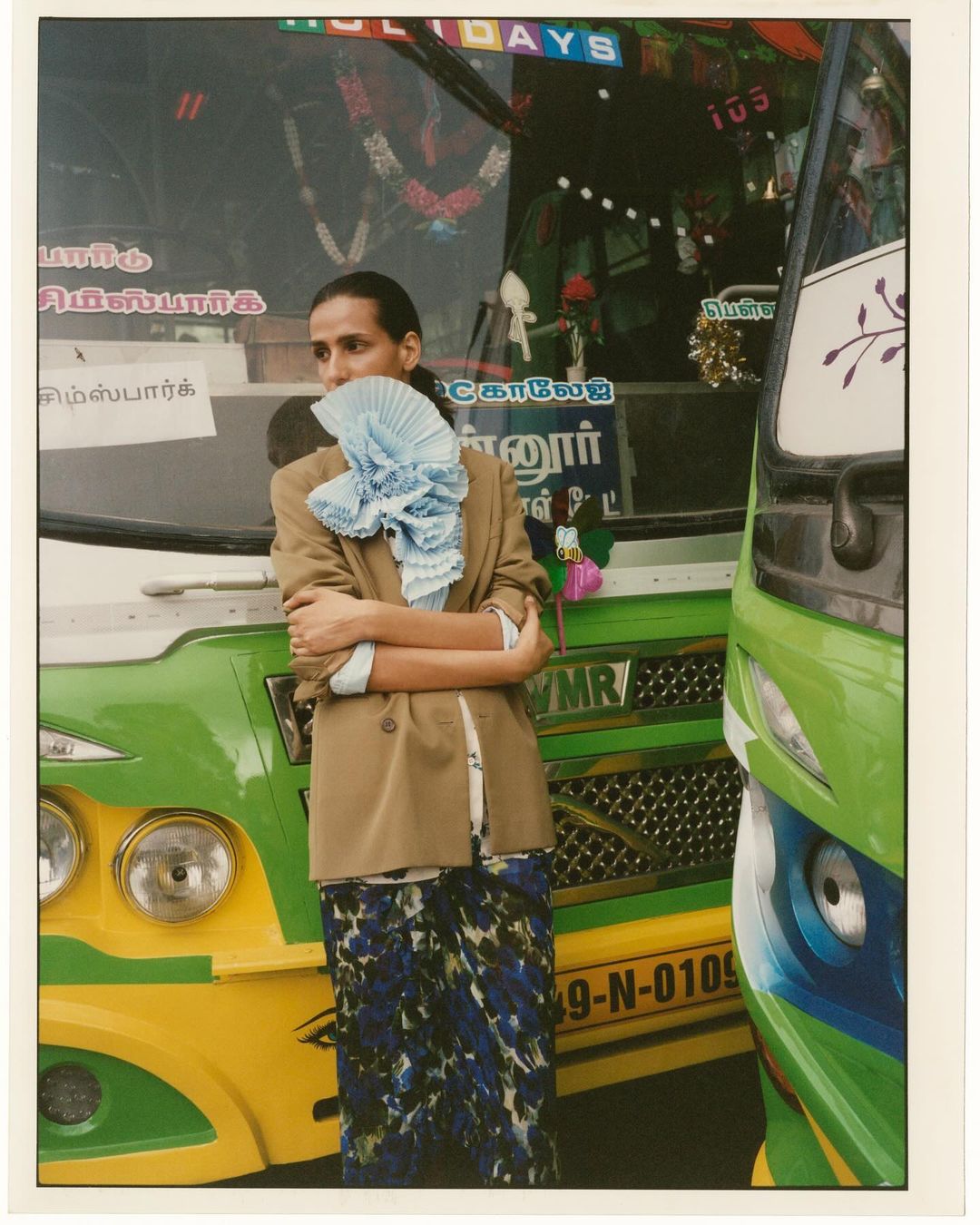 Adult posing in a tan blazer and patterned skirt, standing in front of a green and yellow bus, accessorized with a light blue, ruffled scarf.