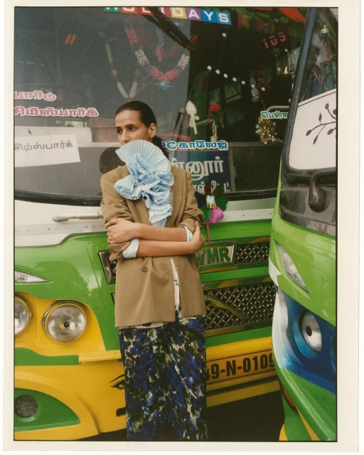 Adult posing in a tan blazer and patterned skirt, standing in front of a green and yellow bus, accessorized with a light blue, ruffled scarf.