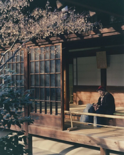 Elderly woman sits on a wooden bench inside a traditional Japanese building with sliding doors and a garden view outside.