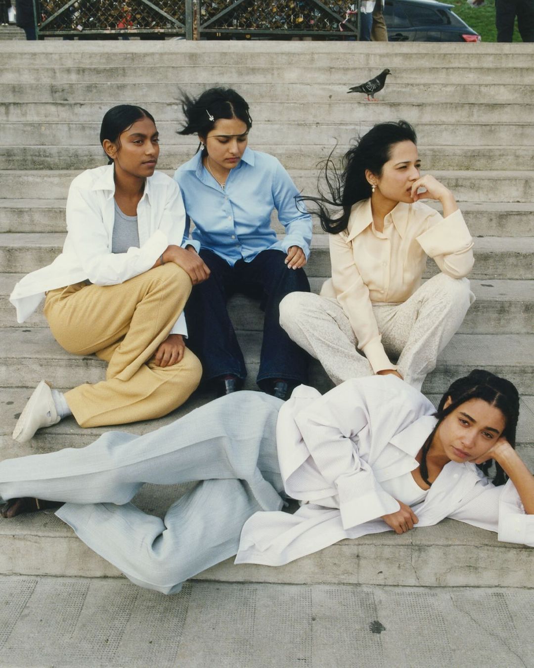 Four young adult women of varying ethnicities sit on concrete steps, wearing casual vintage-style clothes, under daylight in an outdoor setting.