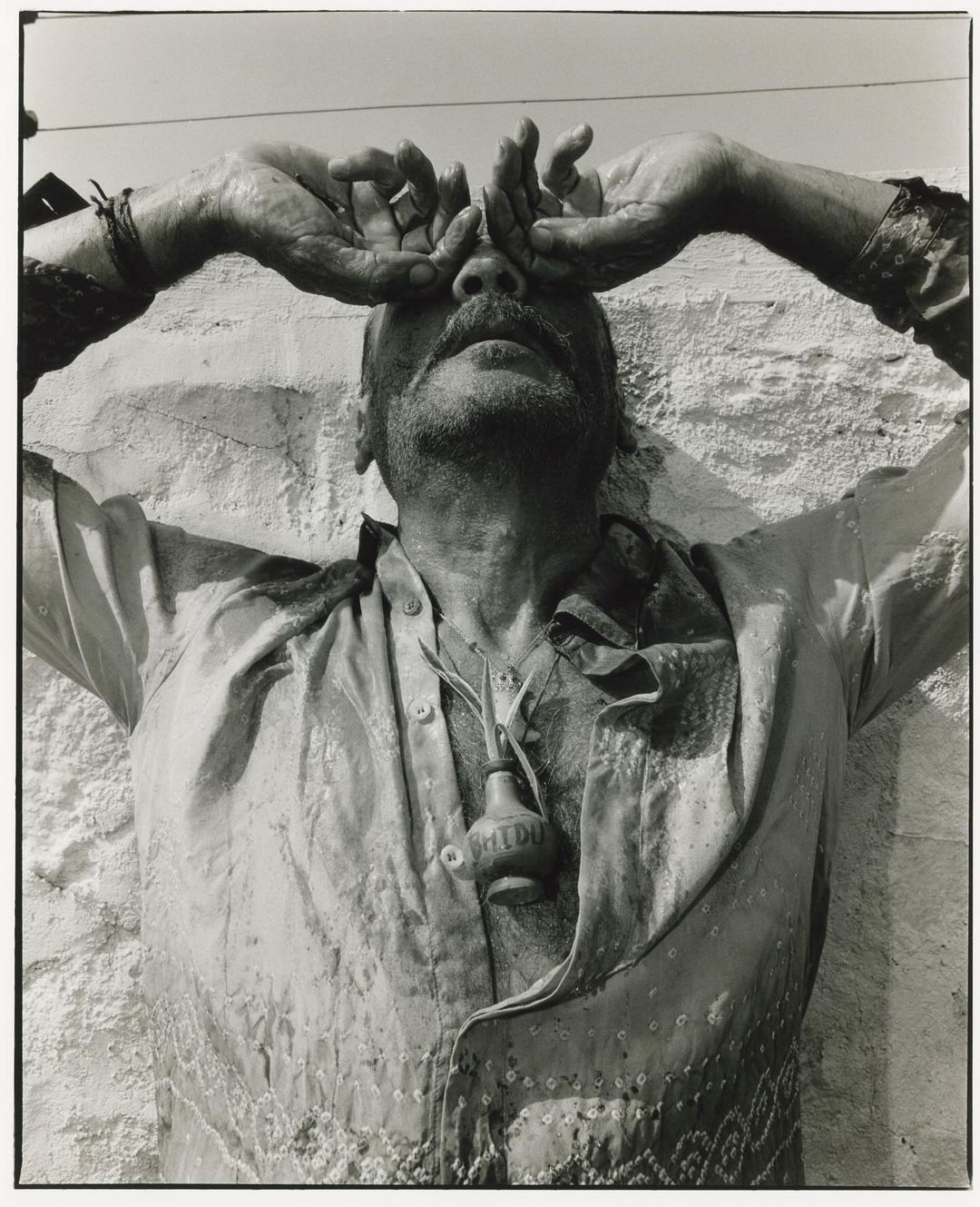 Monochrome portrait of a man with his hands raised to his face, wearing a button-down shirt with a small bottle pendant.