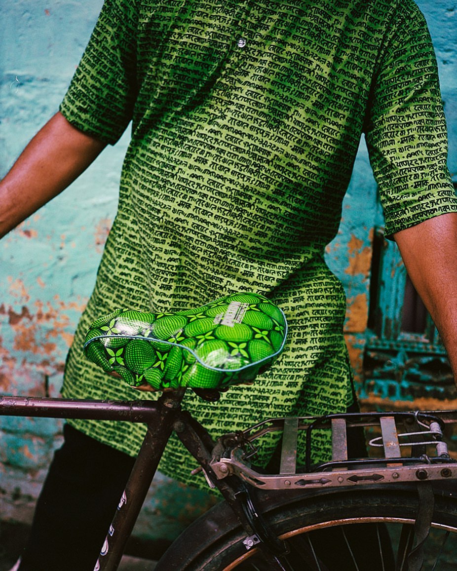 Man in green shirt with text next to bicycle with green patterned seat, standing against a weathered blue wall with window.