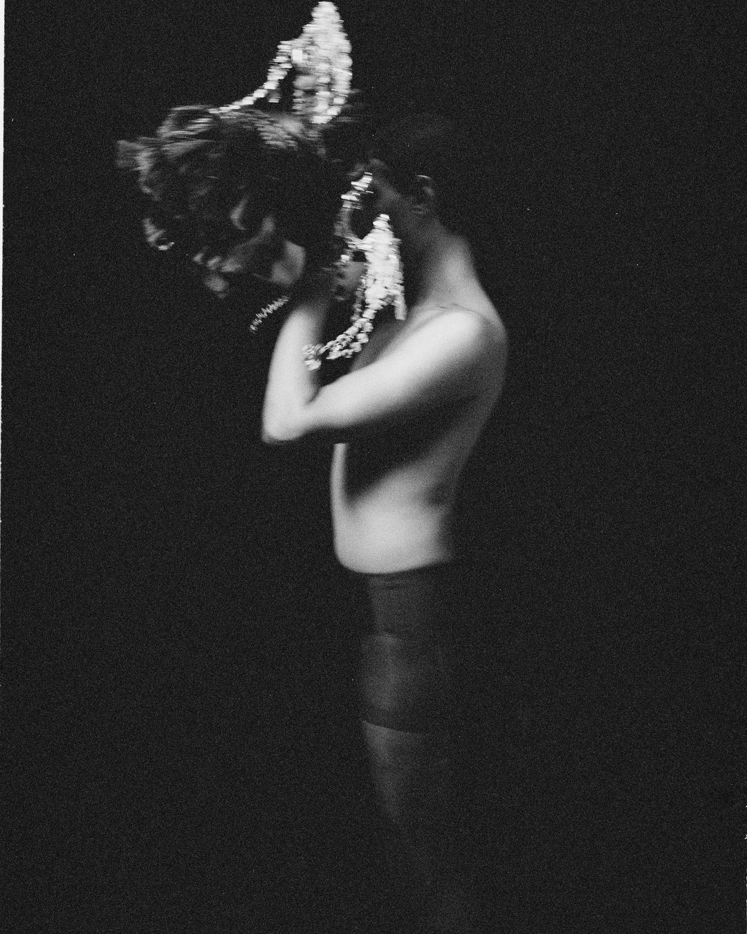 Artistic monochrome photo of a shirtless person wearing a headpiece, facing left with arms crossed, against a dark background.