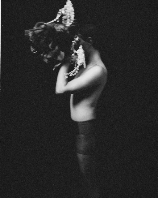 Artistic monochrome photo of a shirtless person wearing a headpiece, facing left with arms crossed, against a dark background.