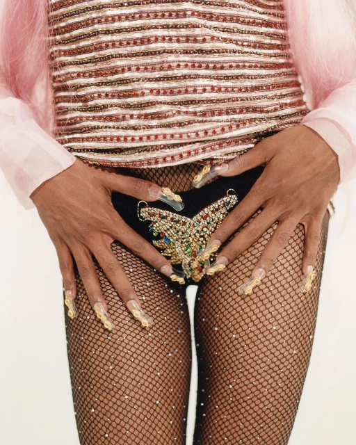 Close-up of a person with decorative nails and rhinestone butterfly on dark shorts over patterned fishnet stockings against a white background.